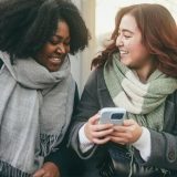 Two young women looking at a smartphone and smiling on a city street, representing mass communication students engaging with digital media