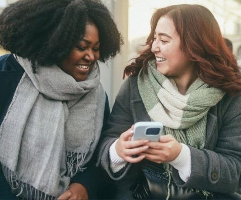 Two young women looking at a smartphone and smiling on a city street, representing mass communication students engaging with digital media