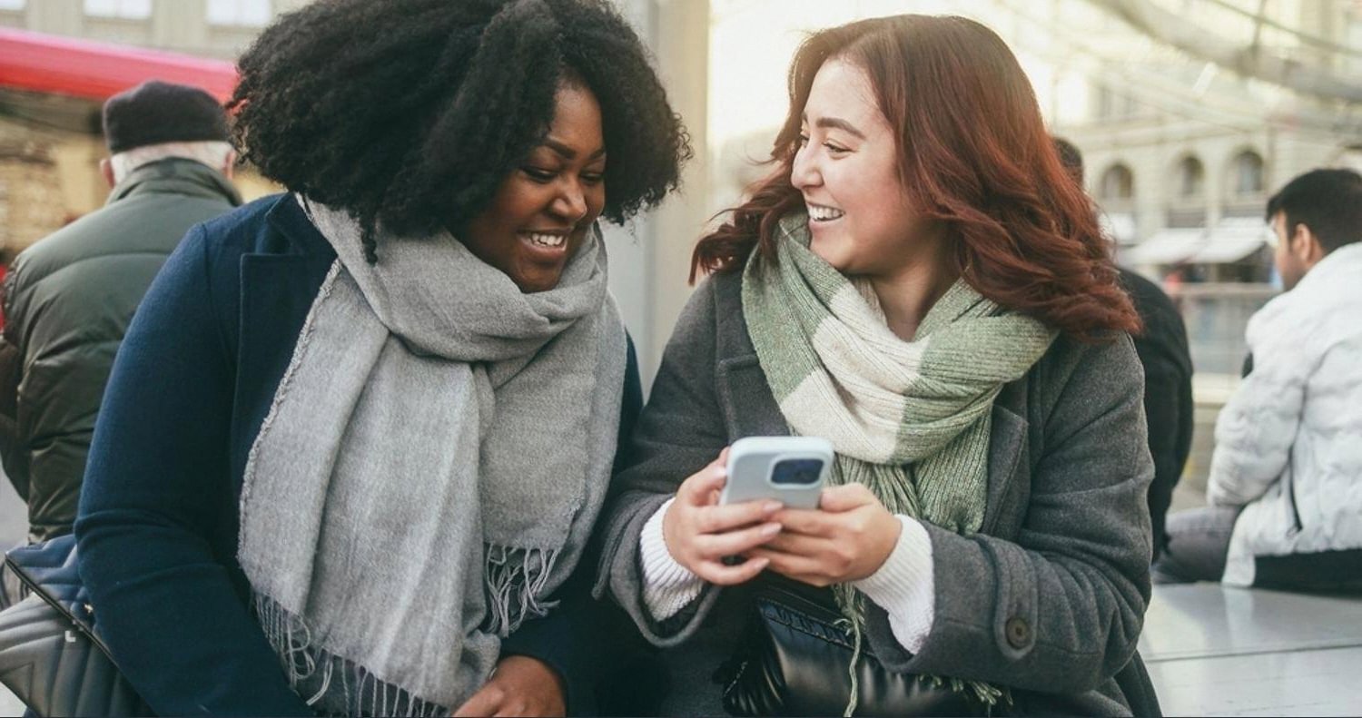 Two young women looking at a smartphone and smiling on a city street, representing mass communication students engaging with digital media
